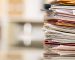 Large stack of file folders, documents, paperwork piled on glass top desk in office.  Bookshelves in background.